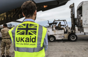 Photo of a man from behind wearing a high visibility jacket with the UK aid logo on it, watching as cargo is loaded onto a military plane.